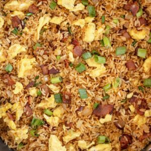 A skillet of Good Morning, Dumbass!—fried rice, scrambled eggs, bacon, and green onions—sits on marble beside a striped towel and a bowl of green onions.