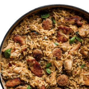 A skillet of Cajun Cluster F@ck with orzo pasta, sliced sausage, chicken, herbs, and fresh basil leaves.