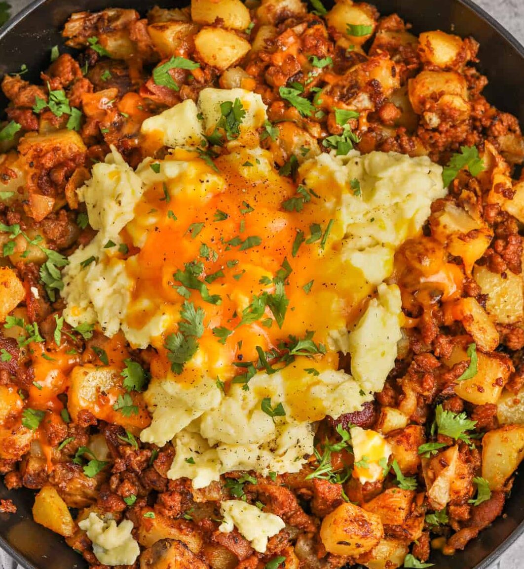 Spank My Hash, And Call Me Sally! is a skillet of diced potatoes, ground meat, cheddar cheese, herbs, with sides of salsa, jalapeños, and chopped vegetables.