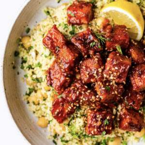 Red Alert Tofu cubes glazed and topped with sesame seeds, served over couscous with chickpeas, herbs, and a lemon wedge.