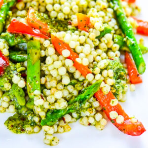 A close-up of pearl couscous mixed with asparagus, red bell pepper strips, and Sweet Heat Chimichurri on a white plate.
