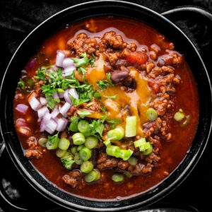 A bowl of chili topped with chopped red onions, green onions, shredded cheese, and fresh parsley.