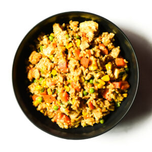 A black bowl filled with rice, diced chicken, carrots, peas, and corn, photographed from above on a white background.