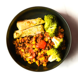 A black bowl containing vegetable stir-fry with corn and tomatoes, steamed broccoli florets, and two slices of seasoned flatbread.