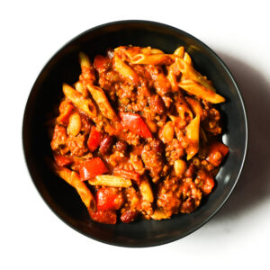 A black bowl filled with pasta, ground meat, beans, tomato sauce, and diced red peppers, photographed from above on a white background.