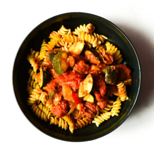 A black bowl filled with rotini pasta topped with a tomato-based sauce, zucchini, and chunks of vegetables, photographed on a white background.
