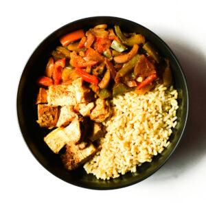 A bowl containing brown rice, grilled tofu cubes, and sautéed bell peppers with onions, photographed from above.