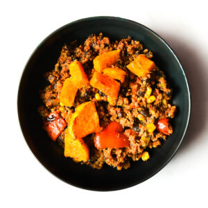 A black bowl filled with a mixture of ground meat, corn, red bell peppers, and topped with chunks of roasted orange squash.