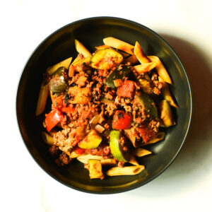 A black bowl filled with penne pasta, ground meat, zucchini, tomatoes, bell peppers, and sauce, viewed from above.