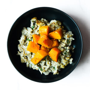 A black bowl containing mashed potatoes with herbs, topped with cubes of roasted butternut squash and sprinkled with black pepper.