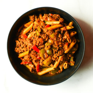 A black bowl filled with penne pasta, ground meat, and chopped bell peppers in a red sauce on a white surface.