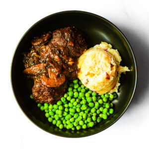 A black bowl containing Salisbury steak with mushroom gravy, a serving of mashed potatoes, and a portion of green peas.