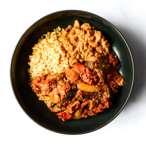 A black bowl containing yellow rice, stewed beans, and a tomato-based beef and vegetable stew, photographed from above on a white background.