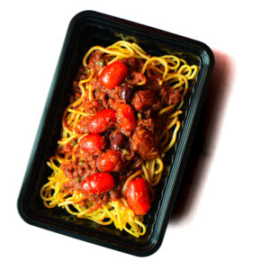 A black plastic tray contains cooked spaghetti topped with tomato sauce, cherry tomatoes, olives, and sausage pieces.