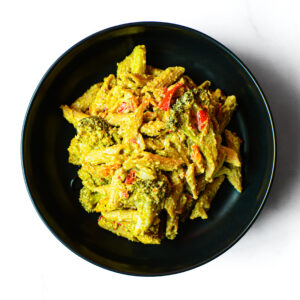 A black bowl filled with penne pasta, broccoli, and red bell peppers in a creamy yellow sauce, viewed from above.