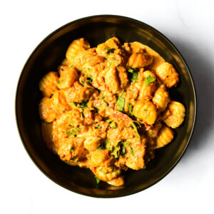 A black bowl filled with creamy gnocchi mixed with pieces of chicken and garnished with fresh herbs, photographed from above.