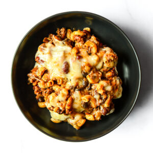 A black bowl filled with macaroni goulash topped with melted cheese, ground meat, beans, and tomato sauce on a white background.