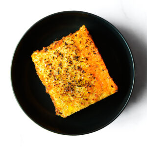 A single slice of baked lasagna with a golden, crisped cheese topping, served on a round black plate.