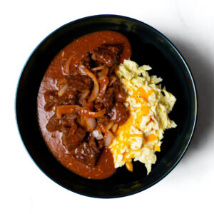 A serving of Meat, Eggs & Consequences: beef stew with onions in red sauce beside cheesy scrambled eggs.