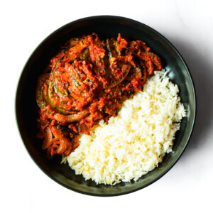A black bowl containing white rice and a tomato-based vegetable stew with sliced zucchini and onions, viewed from above.