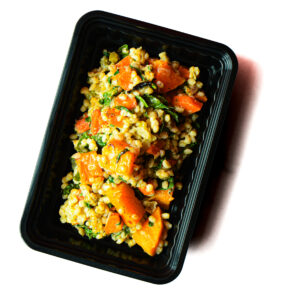 A black plastic container filled with a mixed grain salad featuring chunks of orange squash, leafy greens, and grains.