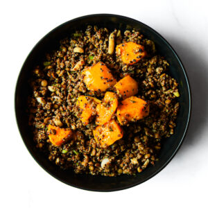 A black bowl filled with a mixture of lentils and beans, topped with chunks of roasted orange squash, on a white background.