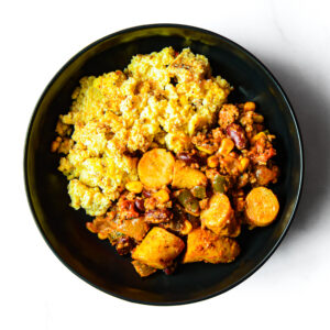 A black bowl containing a serving of scrambled eggs and a vegetable stew with beans, carrots, potatoes, and green peppers, on a white background.