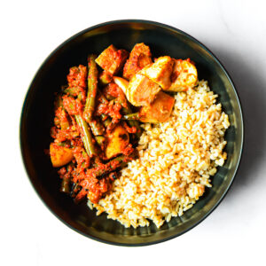 A black bowl containing brown rice on one side and a mixed vegetable curry with tofu in a red sauce on the other side.