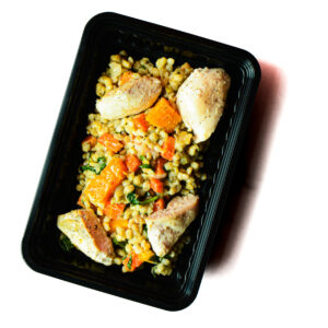A black plastic tray containing cooked chicken breast pieces, roasted vegetables, and a grain or pasta mix.