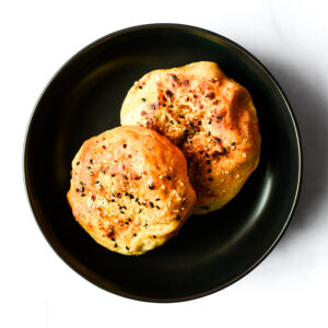 Two round pieces of bread with a golden-brown crust and sprinkled with black sesame seeds, served on a black plate.
