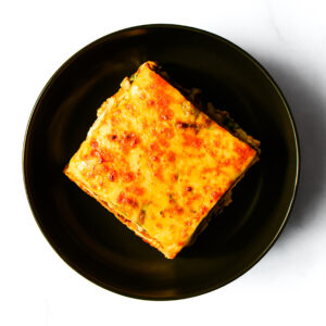 A square slice of This Ain't Buffalo, Clucker! - WL with golden-brown cheese on a round black plate, viewed from above.