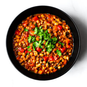 A black bowl filled with bean and vegetable stew, garnished with fresh cilantro, photographed from above on a white surface.