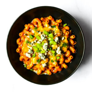 A black bowl filled with macaroni in orange sauce, topped with blue cheese crumbles, chopped celery, and herbs, on a white surface.