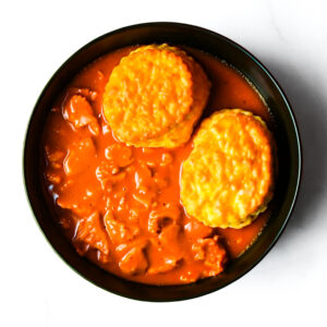A black bowl containing biscuits served over beef stew with a rich orange-brown gravy, viewed from above on a white surface.