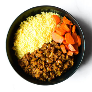 A black bowl containing yellow couscous, cooked sliced carrots, and spiced ground meat with onions, arranged in separate sections.