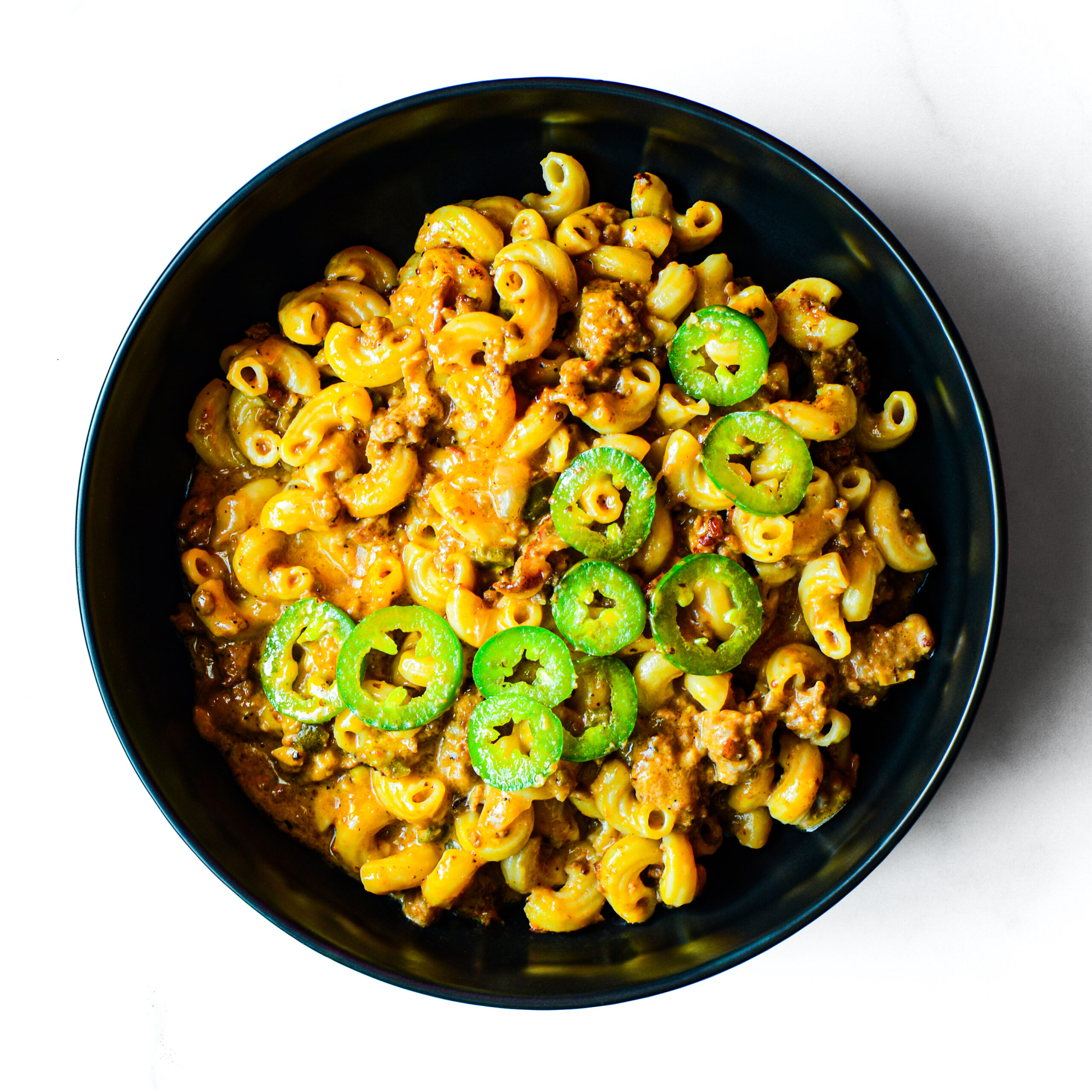 A black bowl filled with macaroni and ground meat in a creamy sauce, topped with sliced green jalapeños, on a white surface.