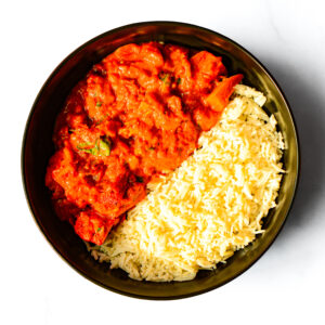 A bowl filled with yellow basmati rice on one side and a red tomato-based curry with visible chunks and herbs on the other side.