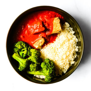 A bowl containing white rice, steamed broccoli, and tofu in red tomato-based sauce, arranged on a white surface.