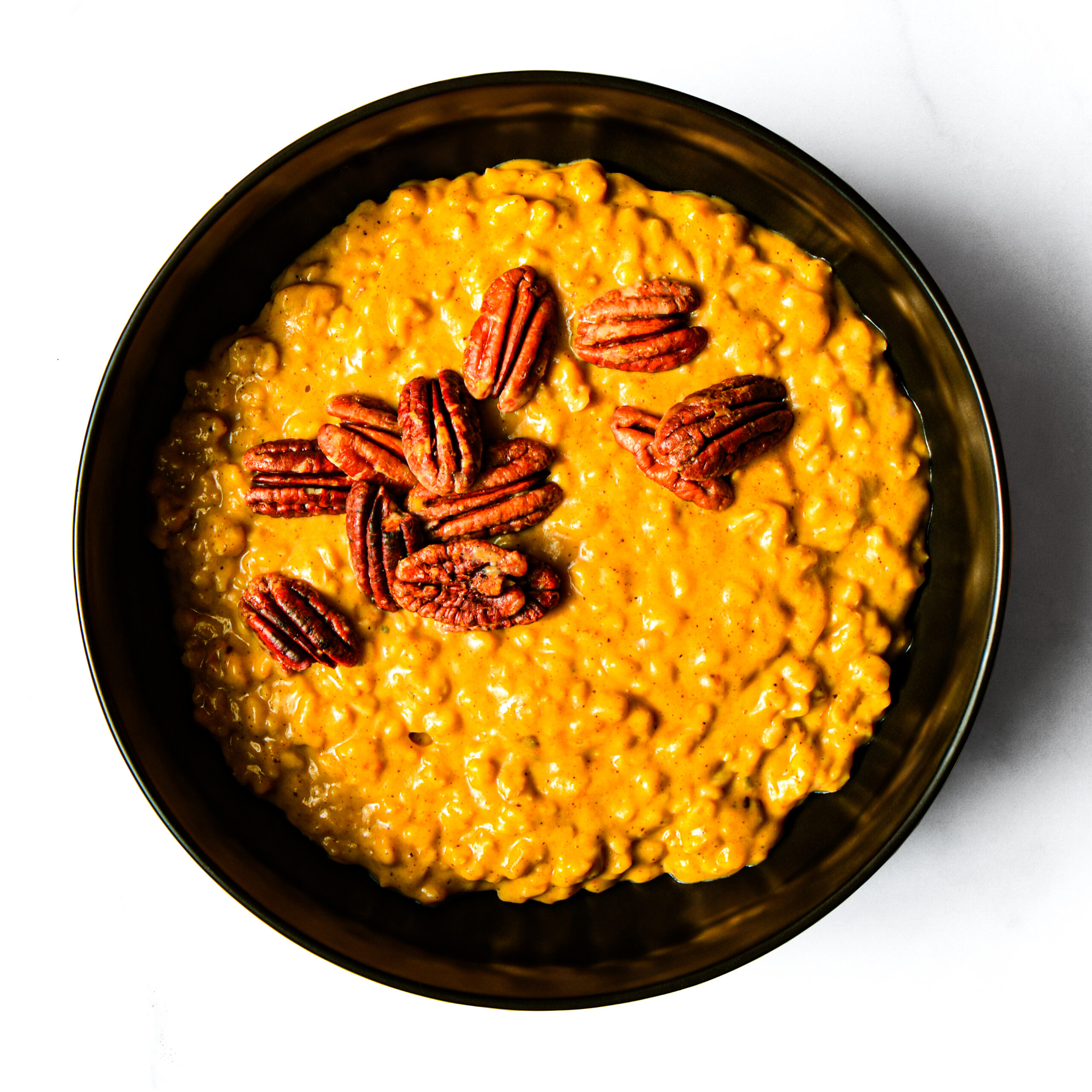 A black bowl filled with orange pumpkin oatmeal topped with whole pecans, viewed from above on a white background.