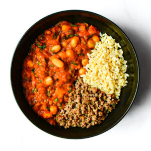 A black bowl containing cooked ground meat, yellow rice, and chickpeas in tomato sauce, arranged in separate sections.