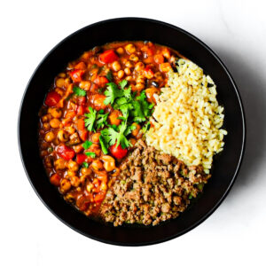 A black bowl containing chili with chickpeas and red peppers, a portion of rice, and ground beef, garnished with fresh cilantro.