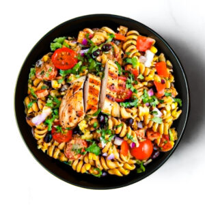 A black bowl filled with rotini pasta salad, grilled chicken slices, cherry tomatoes, black beans, corn, red onion, and chopped herbs.