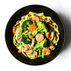 A black bowl filled with fettuccine pasta, broccoli florets, sliced carrots, mushrooms, and green peas in a creamy sauce, viewed from above.