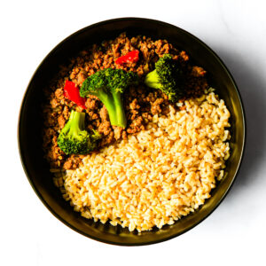 A black bowl containing cooked ground meat with broccoli and red bell pepper on one side, and a serving of brown rice on the other.