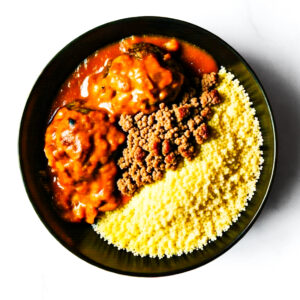A black bowl containing yellow couscous, minced meat, and two meatballs in red sauce on a white background.