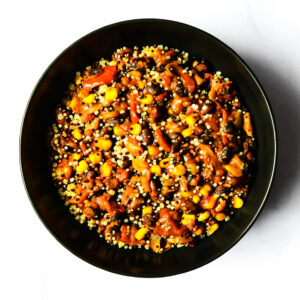 A black bowl filled with a mixed dish of beans, corn, tomato, and grains, photographed from above on a white background.