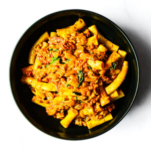A black bowl filled with tube-shaped pasta in a creamy tomato sauce with visible herbs and pieces of vegetables.