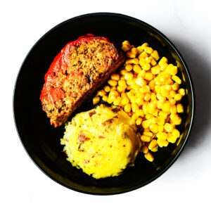 A black plate with a slice of meatloaf, a serving of corn, and a scoop of mashed potatoes.