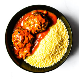 A black bowl containing two meatballs in tomato sauce on one side and a serving of plain couscous on the other.