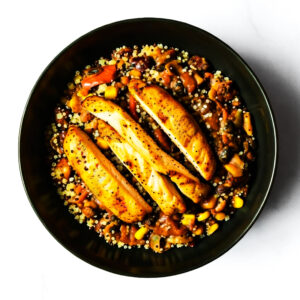 A black bowl with grilled chicken breast slices on top of a mixture of quinoa, corn, beans, and diced vegetables.